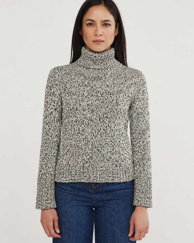 A woman modeling a long-sleeve knit turtleneck sweater with a black and white marled pattern, paired with blue jeans, against a plain light gray studio background.