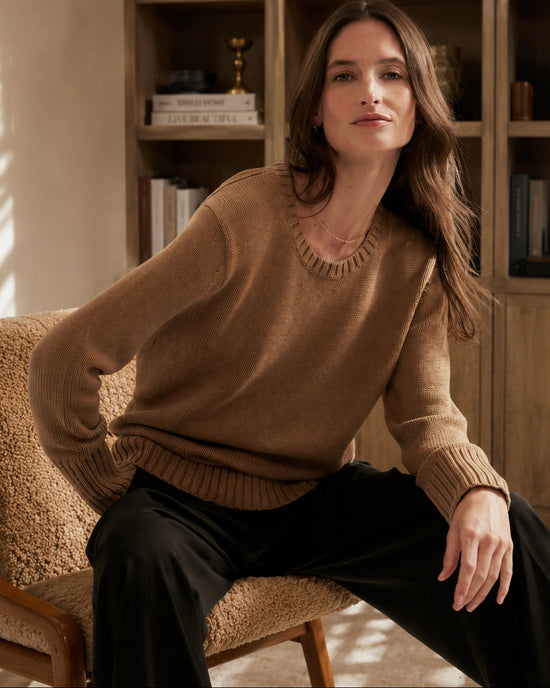 A woman sits on a textured armchair in a warm, wood-paneled room, wearing a camel brown crewneck knit sweater and black trousers.