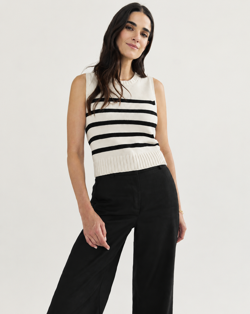 A woman modeling a sleeveless knit top with horizontal black and off-white stripes, paired with black wide-leg pants, against a plain light gray studio background.