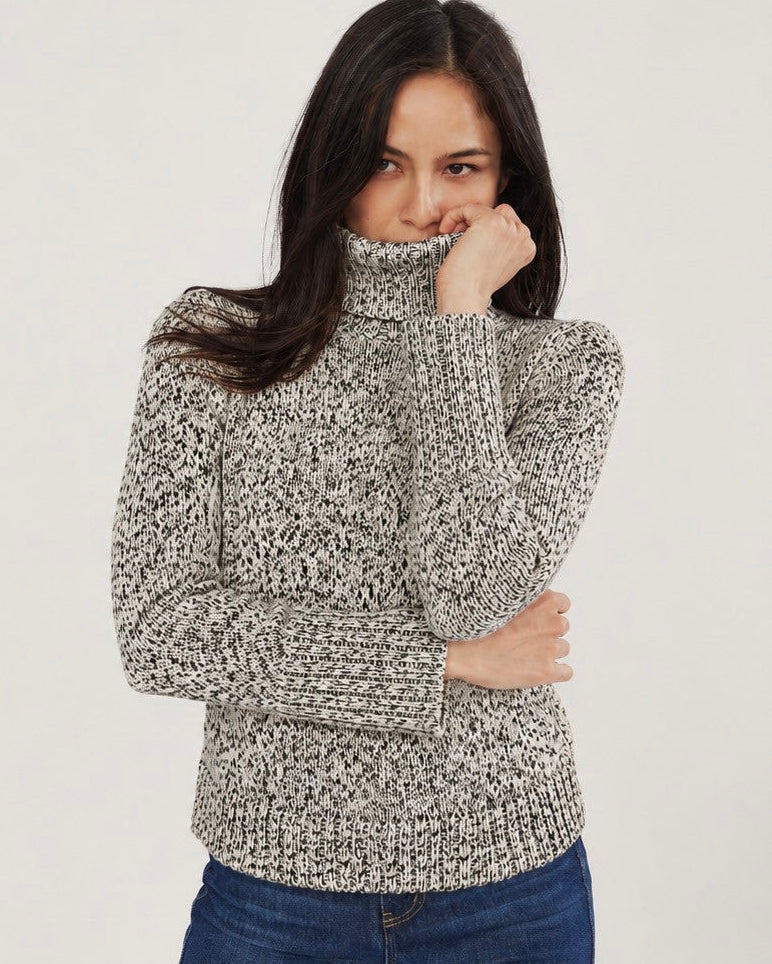 A woman wearing a chunky knit turtleneck sweater in a heathered gray and white marled pattern, paired with blue jeans, against a plain light gray background.